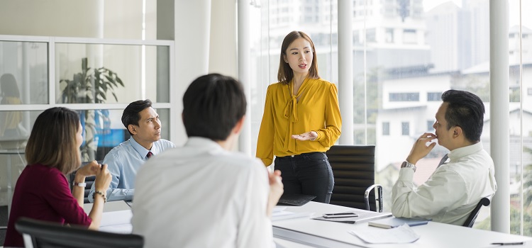 Five office workers in a circle meeting room with one of them standing up, while the others are looking at them